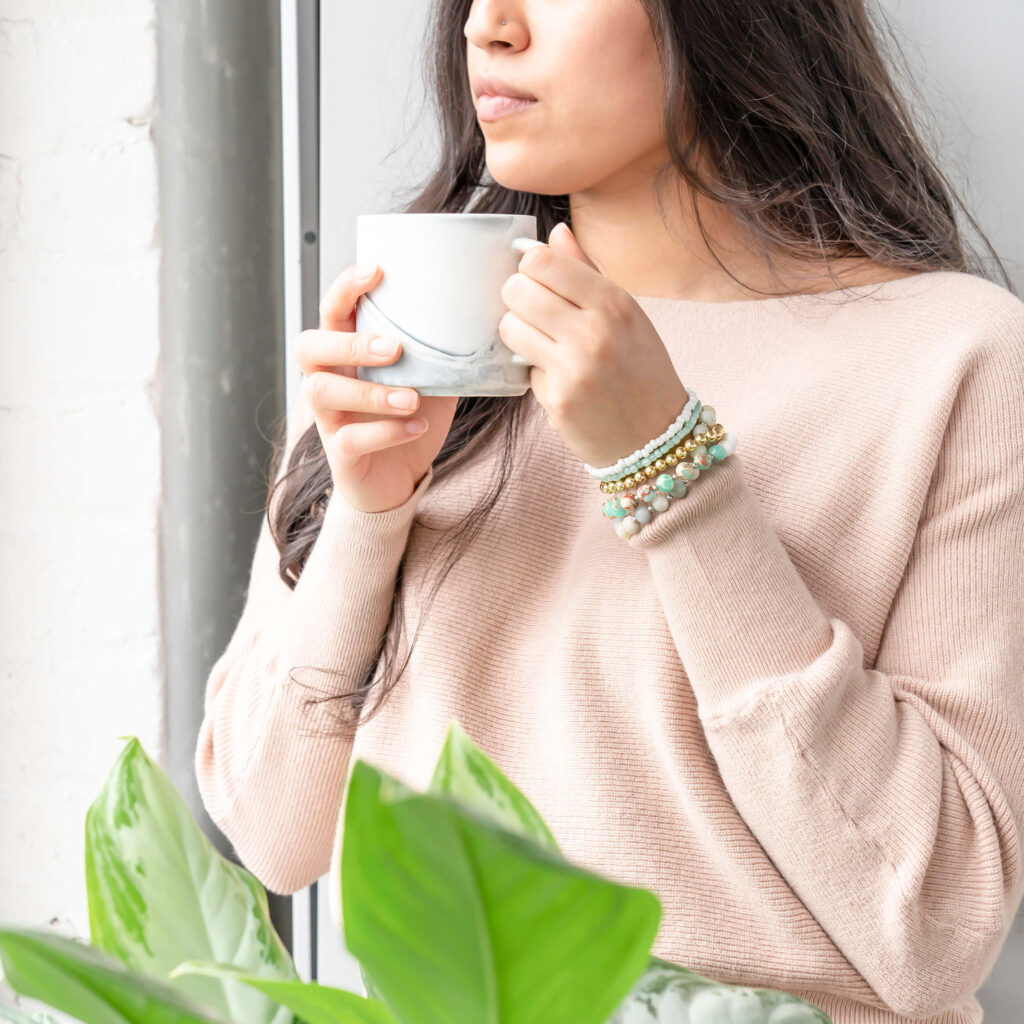 A woman in pastel pink sweater is holding a mug, looking out the window as she sips on calming herbal tea. She is setting herself up for good sleep.