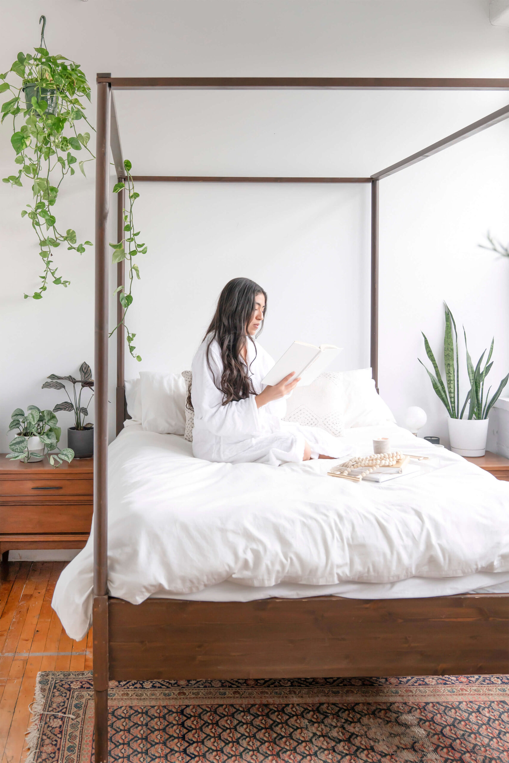 A woman all in white on crisp white-linen bed, reading a book beautiful and serene looking room with a big wooden framed bed. This images conveys peace and tranquility.