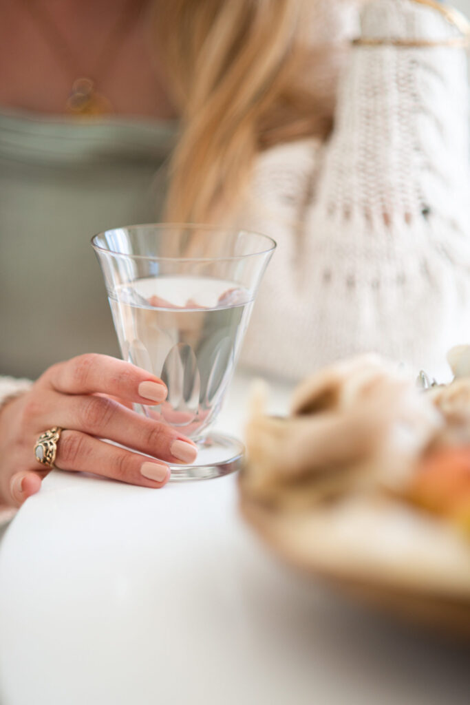 Blonde woman holding a glass of water. The picture is blurry and zoomed in on the glass of water. Burgundy scrunchy and sports bra, ear pods and a glass reusable water bottle filled with iced-water. Optimal hydration and knowing what and when to drink are key to well-being and performance.