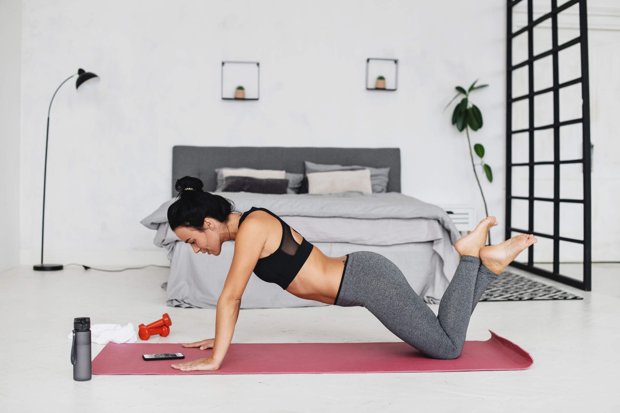 Young woman practicing yoga at home in her bedroom; She has a pink yoga mat and it using her phone to guide her in her home practice