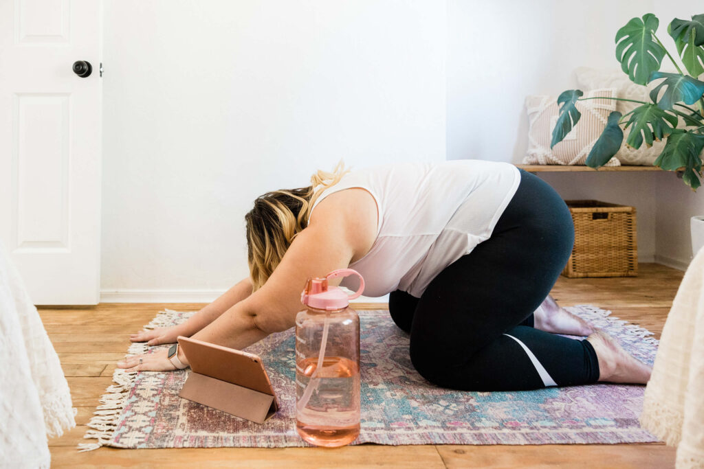 Larger fair skinned woman practising child's pose at home with her yoga routine on her ipad guiding her in her yoga home practice