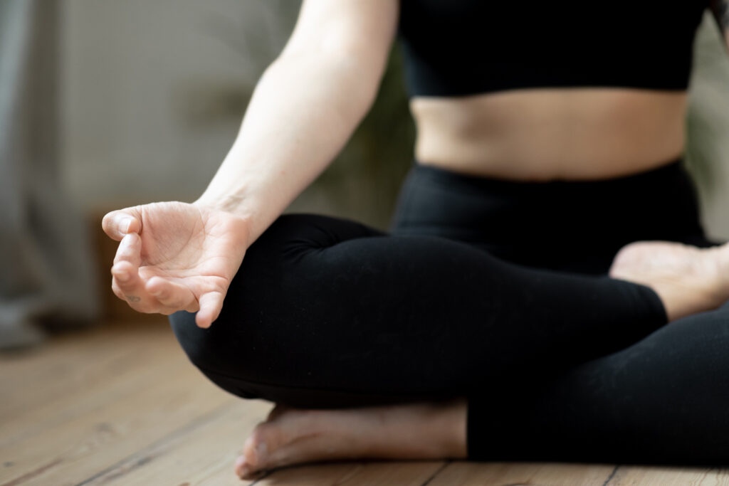 Girl meditating with a hand mudra, wearing a black sports bra and black leggings, neutral tones. This images is expressing the zen vibes you get from meditating or private yoga classes.