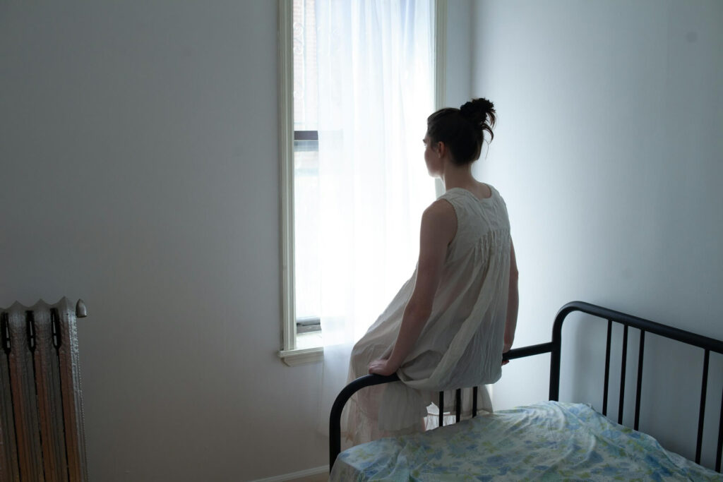 young womand in white nightgown in white room mindlessly staring out a window