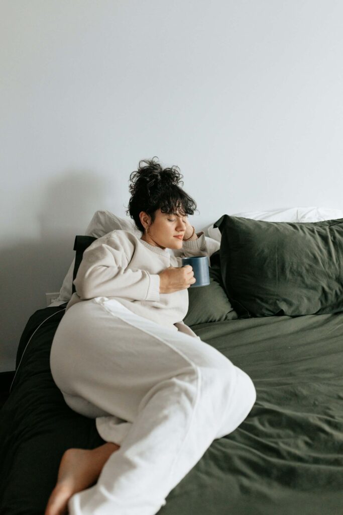 woman holding a hot beverage and resting on her bed.